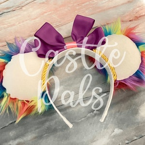 May include: Rainbow-coloured faux fur ear headband with a white felt base and a purple satin bow. The headband is gold and white.
