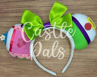 May include: A headband featuring two padded ears with a large lime green satin bow. One ear is white with a pink square, a red heart, and lace trim. The other ear is white, lime green, and purple with a red circle and a yellow tag reading LIGHTYEAR.