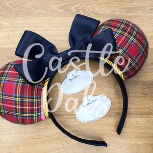 May include: Red and black plaid Minnie Mouse ears with a black satin bow and gold rope trim. The ears come with two blank name tags, one that says "Disneyland" and one that says "Disney World".