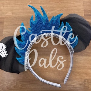 May include: Blue and black glitter Minnie Mouse ears with a silver headband and a blue flame design. The ears have black tulle and a white cupcake design on one ear. The headband has the text "Castle Pals" on it.
