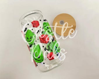 May include: A clear glass tumbler with a green and red design featuring a cartoon character with a green face and red dice. The tumbler has a bamboo lid.