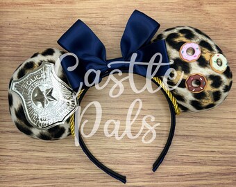 May include: A black headband with leopard print ears. One ear features a silver police badge, the other has three donut embellishments. A navy blue bow sits atop the headband. The text "Castle Pals" is visible.