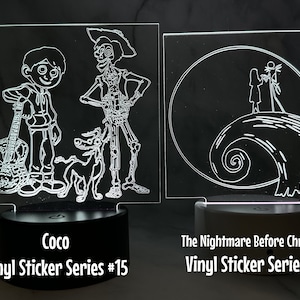 Puede incluir: Dos letreros acrílicos iluminados con dibujos de líneas blancas. El letrero de la izquierda presenta personajes de la película Coco, incluidos Miguel, Héctor y Dante. El letrero de la derecha presenta a Jack y Sally de la película El extraño mundo de Jack. Ambos letreros tienen una base negra y el texto "Vinyl Sticker Series" con un número.