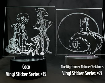 May include: Two illuminated acrylic signs with white line drawings. The left sign features characters from the movie Coco, including Miguel, Hector, and Dante. The right sign features Jack and Sally from the movie The Nightmare Before Christmas. Both signs have a black base and the text "Vinyl Sticker Series" with a number.