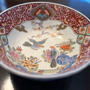 May include: A white ceramic bowl with a red, gold, and blue floral design. The bowl features two birds and a floral pattern.