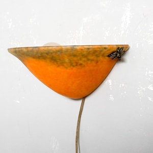 Op de afbeelding: Een oranje en bruine wandlamp met een uniek, gebogen ontwerp. De lamp is bevestigd aan een witte muur en heeft een lange, dunne draad die naar beneden loopt. De lamp heeft een decoratief element aan de zijkant.