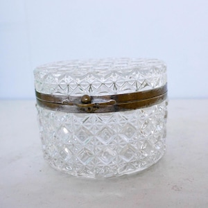 Op de afbeelding: Een heldere, ronde glazen sieradendoos met een ruitpatroon. De doos heeft een scharnierend deksel en is omlijst met een aangetaste messing metalen band. De doos staat op een wit oppervlak.