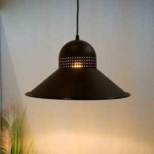 Op de afbeelding: Een zwarte metalen hanglamp met een brede, platte kap en een kleinere, geperforeerde bovenkant. De lamp is aan en geeft een warm licht.