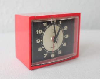 May include: A red alarm clock with a black face and white numbers. The clock has a clear plastic cover over the face.