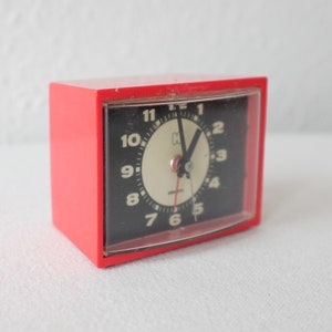 Può includere: Una sveglia rossa con quadrante nero e numeri bianchi. L'orologio ha una copertura in plastica trasparente sul quadrante.