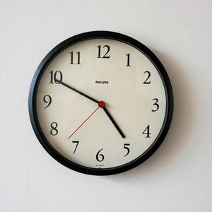 Puede incluir: Un reloj de pared redondo con marco negro y esfera color hueso. El reloj presenta números arábigos negros del 1 al 12, el nombre de la marca Philips en texto negro, manecillas negras para horas y minutos, y un segundero rojo muy fino.