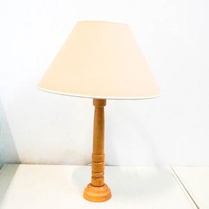 Op de afbeelding: Een houten tafellamp met een beige lampenkap van stof. De lamp heeft een eenvoudig ontwerp met een cilindrische voet en een ronde kap.