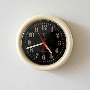 Puede incluir: Un reloj de pared redondo con un marco grueso de color crema y una esfera negra. El reloj presenta números arábigos blancos, manecillas blancas para las horas y los minutos, y un segundero de color naranja brillante. Pequeños puntos naranjas marcan los minutos.