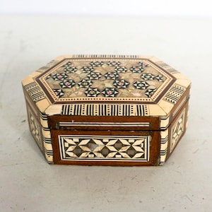 May include: An octagonal wooden box with inlaid mother-of-pearl and geometric designs. The box has a dark brown wood base with decorative patterns on the sides and lid. The lid is adorned with floral and geometric patterns in shades of white, pink, and black.