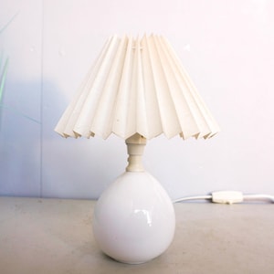 May include: A small, white table lamp with a pleated, off-white lampshade. The lamp has a rounded base and a slender neck. The lamp is on a light gray surface, with green plants in the background. The lamp is plugged into a wall outlet.