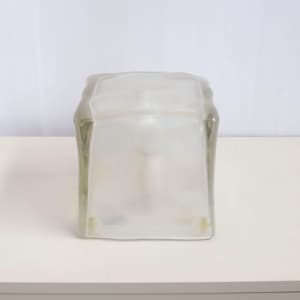 May include: A clear, frosted glass jar with a rectangular shape. The jar has a slightly curved top and is sitting on a white surface. The glass has a frosted appearance, obscuring the contents within.