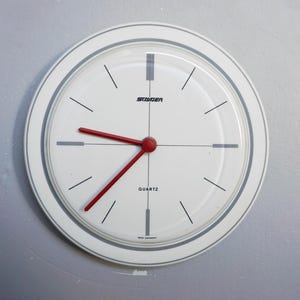 Op de afbeelding: Witte quartz wandklok met een grijze rand en rode wijzers. De wijzerplaat heeft zwarte lijnen die de uren markeren en het woord "STAIGER" bovenaan. Het woord "QUARTZ" staat onderaan gedrukt.