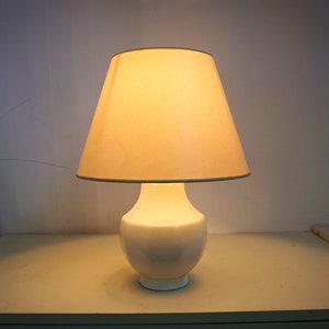 Op de afbeelding: Een witte keramische tafellamp met een witte lampenkap van stof. De lamp is aan en het licht schijnt.