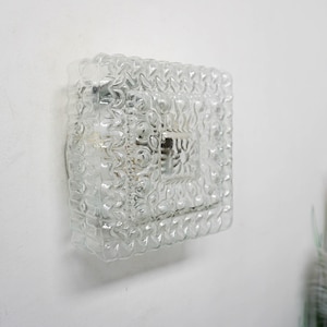 May include: A clear glass wall sconce with a textured, geometric pattern. The sconce is mounted on a white wall.