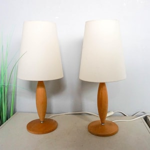 Op de afbeelding: Twee bijpassende tafellampen met crèmekleurige kappen en houten voetstukken. De lampen hebben een klassiek ontwerp, met een ronde voet en een slanke, zandlopervormige steel. De kappen zijn in een zachte, neutrale kleur.