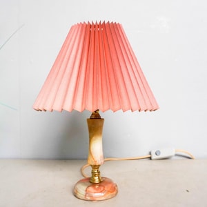 Op de afbeelding: Een vintage tafellamp met een geplooide roze lampenkap. De lamp heeft een houten voet met messing accenten en een marmerachtige bodem. De lamp staat op een lichtgekleurd oppervlak.