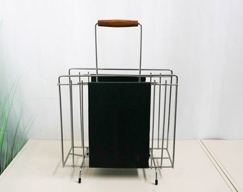 Puede incluir: Un revistero de metal plateado con un asa de madera y insertos para revistas negros. El revistero está sobre una superficie blanca.