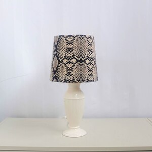 May include: A white ceramic table lamp with a snakeskin patterned lampshade. The lampshade is black and beige with a geometric pattern.