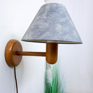May include: A wooden wall sconce with a grey fabric lampshade. The sconce has a round base and a long arm that extends out to the lampshade.