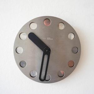 Peut inclure: Une horloge murale circulaire au fini métallique brossé dotée de douze découpes circulaires sur le pourtour. L'horloge possède deux aiguilles noires, l'une pleine et l'autre avec une fente allongée, positionnées pour indiquer l'heure sur le cadran.
