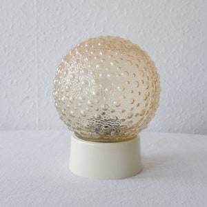 Op de afbeelding: Een vintage tafellamp met een getextureerde, bolvormige glazen kap in een warme amberkleur. De kap staat op een crèmekleurige, cilindrische voet. De lamp is ontworpen om zachte, sfeervolle verlichting te bieden.