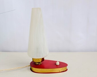 Könnte beinhalten: Eine rote und goldene Tischlampe mit einem weißen, gefalteten Lampenschirm. Die Lampe hat einen runden Sockel mit goldener Verzierung und einen kleinen weißen Schalter.