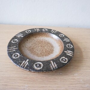 Op de afbeelding: Een ronde keramische schaal met een gespikkeld beige centrum en een donkergrijze rand. De rand is versierd met een herhalend patroon van ingesneden cirkels en groepjes van drie verticale lijnen, wat een textuurcontrast geeft met de gladde kom.