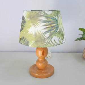 Op de afbeelding: Een houten tafellamp met een witte lampenkap met een tropisch bladmotief. De lamp heeft een ronde voet en een ontwerp met dubbele lampen.