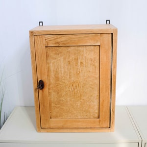 Op de afbeelding: Een licht houten wandkast met een enkele deur en een metalen sluiting. De kast heeft een eenvoudig ontwerp en is gemaakt van natuurlijk hout.