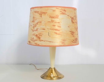 May include: A vintage table lamp with a white ceramic base and a gold metal bottom. The lampshade is a light brown with a peach trim and a marbled pattern.