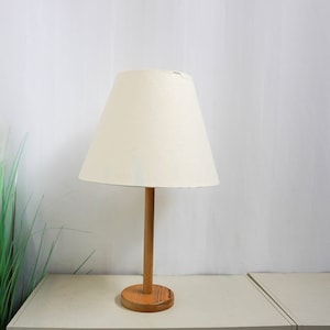 Op de afbeelding: Een witte lampenkap met een houten voet staat op een witte tafel. De lampenkap is gemaakt van stof en heeft een eenvoudig ontwerp. De voet is gemaakt van hout en heeft een natuurlijke afwerking.