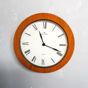 May include: Round wall clock with a wooden frame and a white face. The clock features black Roman numerals, black hands, and the word "QUARTZ" at the bottom. The brand name "JUNGHANS" is printed at the top.