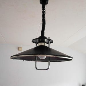 Könnte beinhalten: Eine schwarze, kegelförmige Pendelleuchte mit perforiertem Schirm und silberner Zierleiste. Das Licht hängt an einem schwarzen, flexiblen Kabel von der Decke. Eine einzelne Glühbirne ist im Schirm sichtbar. Modernes Design.
