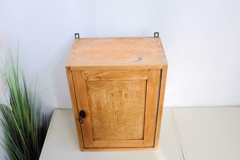 Op de afbeelding: Een klein houten kastje met een enkele deur en een sleutelgat. Het kastje heeft metalen scharnieren en is in een lichte bruine kleur geverfd.