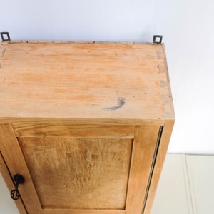 Op de afbeelding: Een klein houten kastje met een enkele deur en een sleutelgat. Het kastje is lichtbruin en heeft een eenvoudig ontwerp. Het is aan een muur bevestigd met twee metalen beugels.