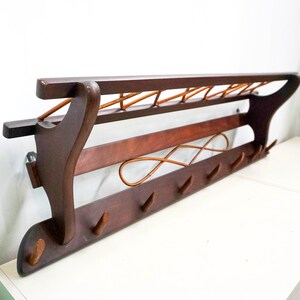 May include: A dark brown wooden wall-mounted coat rack with a shelf and multiple hooks. The rack has a decorative woven design on the shelf.