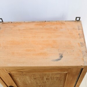 Op de afbeelding: Een houten kast met een enkele deur en twee metalen scharnieren. De kast is gemaakt van licht hout en heeft een natuurlijke afwerking.