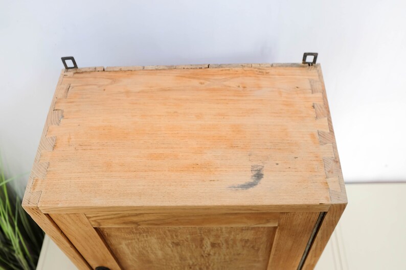 Op de afbeelding: Een houten kast met een enkele deur en twee metalen scharnieren. De kast is gemaakt van licht hout en heeft een natuurlijke afwerking.