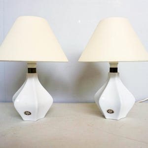 Könnte beinhalten: Zwei weiße Tischlampen mit cremefarbenen Schirmen. Die Lampen haben ein einzigartiges, geometrisches Basisdesign und einen dunkelbraunen Akzent. Sie stehen auf einer weißen Oberfläche vor einer hellgrauen Wand.
