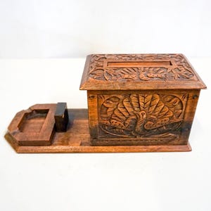 Può includere: Una scatola di legno con un intricato design intagliato di pavoni. La scatola ha un coperchio incernierato e un piccolo vassoio sul lato. La scatola è in legno scuro e ha una finitura naturale.
