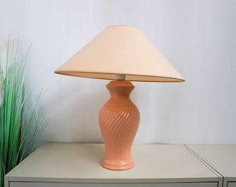 May include: A peach-coloured ceramic table lamp with a white fabric lampshade. The lamp has a swirl design and a round base.