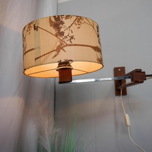 May include: A vintage wall-mounted lamp with a beige lampshade featuring a brown botanical pattern. The lamp has a wooden base and a chrome arm. The lamp is plugged into a wall outlet.