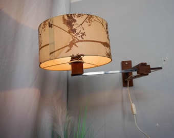 May include: A vintage wall-mounted lamp with a beige lampshade featuring a brown botanical pattern. The lamp has a wooden base and a chrome arm. The lamp is plugged into a wall outlet.