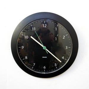 以下が含まれることがあります： 黒い文字盤と白い数字の丸い黒い壁掛け時計。 時計には白い分針と時針、緑色の秒針が付いています。 ブランド名「PHILIPS」が文字盤の下部に印刷されています。