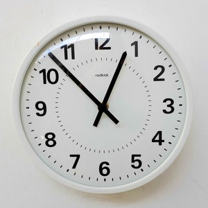 May include: A white analog clock with black hands and black numbers on a white background. The clock face has a small black logo that says "nedok" in the center.